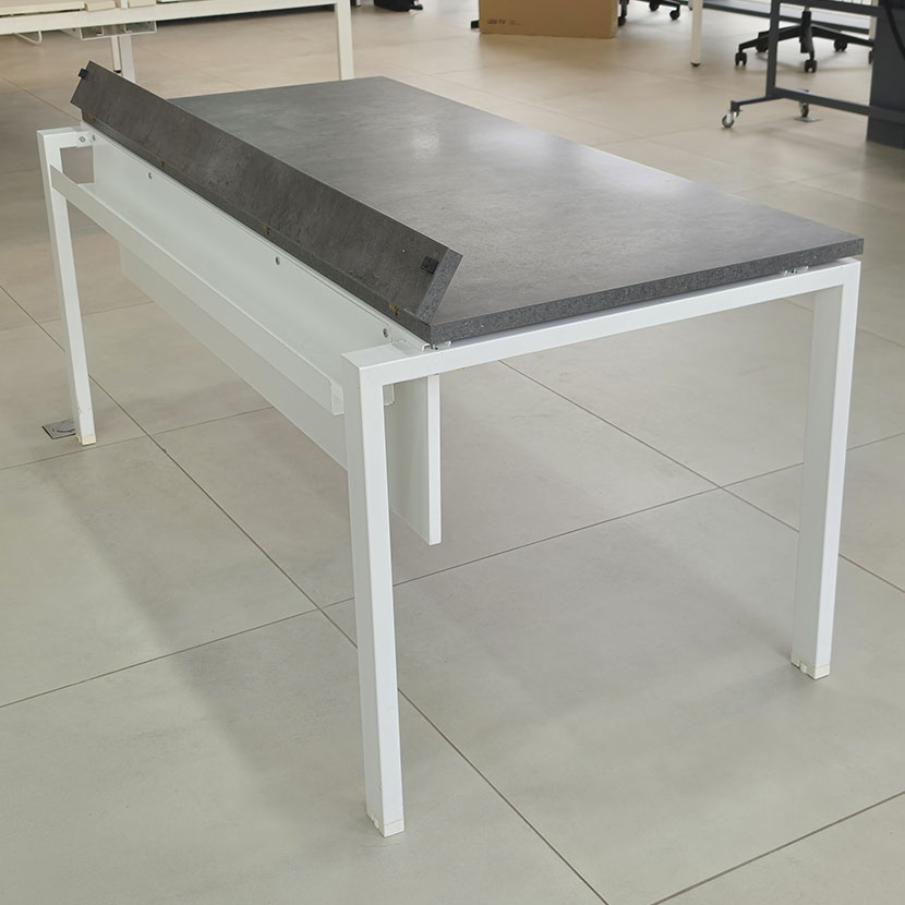 Vue de l'avant à l'ouverture du passage des câble d'une table de bureau professionnelle avec plateau gris béton, piètement métallique blanc et voile de fond.